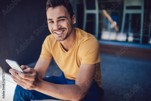 Joyful guy sitting next to building and using mobile phone