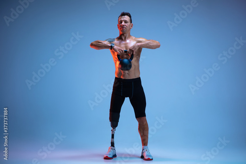 Athlete with disabilities or amputee isolated on blue studio background. Professional male sportsman with leg prosthesis training with weights in neon. Disabled sport and overcoming, wellness concept.