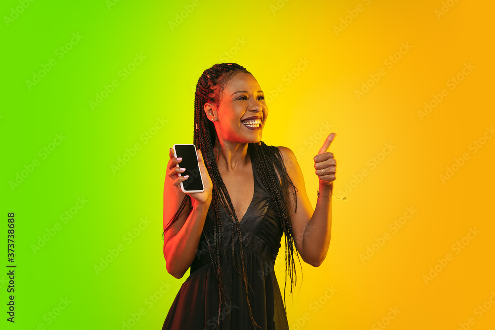 Holding phone with thumb up. Portrait of young woman in neon light on gradient backgound. The human emotions, black friday, cyber monday, purchases, sales, finance concept. Trendy colors. Holding phone with thumb up. Portrait of young woman in neon light on gradient backgound. The human emotions, black friday, cyber monday, purchases, sales, finance concept. Trendy colors.
