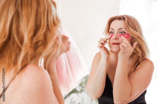 A beautiful young woman takes out pink patches from a package and applies them under her eyes  standing in front of a mirror. Home care concept