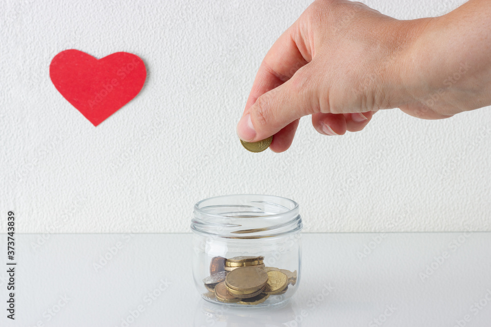 Hand putting coin in a jar on white background with red heart symbol. Saving money or donation concept. Hand putting coin in a jar on white background with red heart symbol. Saving money or donation concept.