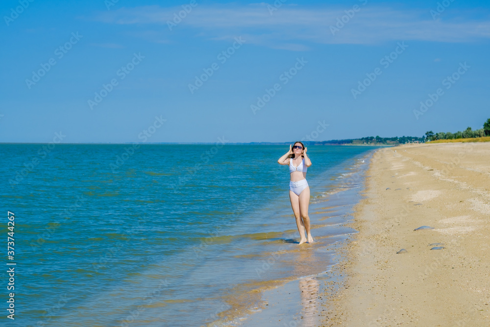 A lonely young girl in a swimsuit and sunglasses stands on the sand near the surf on an empty sea beach. Bright sunny summer day. Calm sea and deep blue sky. Vacation paradise. Copy space. 
