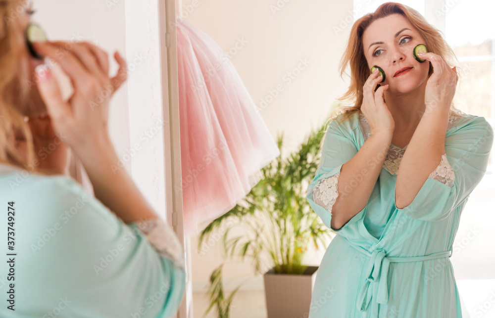 A beautiful woman with a dressing gown and cucumber slices on her face stands in front of the mirror. Home skin care and natural cosmetics concept A beautiful woman with a dressing gown and cucumber slices on her face stands in front of the mirror. Home skin care and natural cosmetics concept