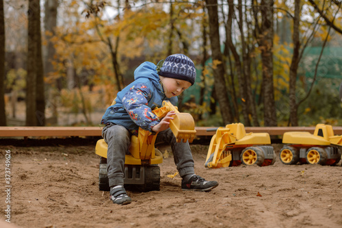 cute little caucasian boy wearing blue warm jacket sitting on a big toy excavaton on playgrownd in autumn