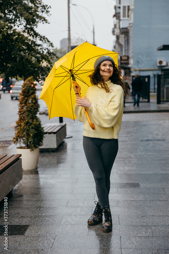 Charming young curly  woman use yellow umbrella at the street of megapolis city in rainy day