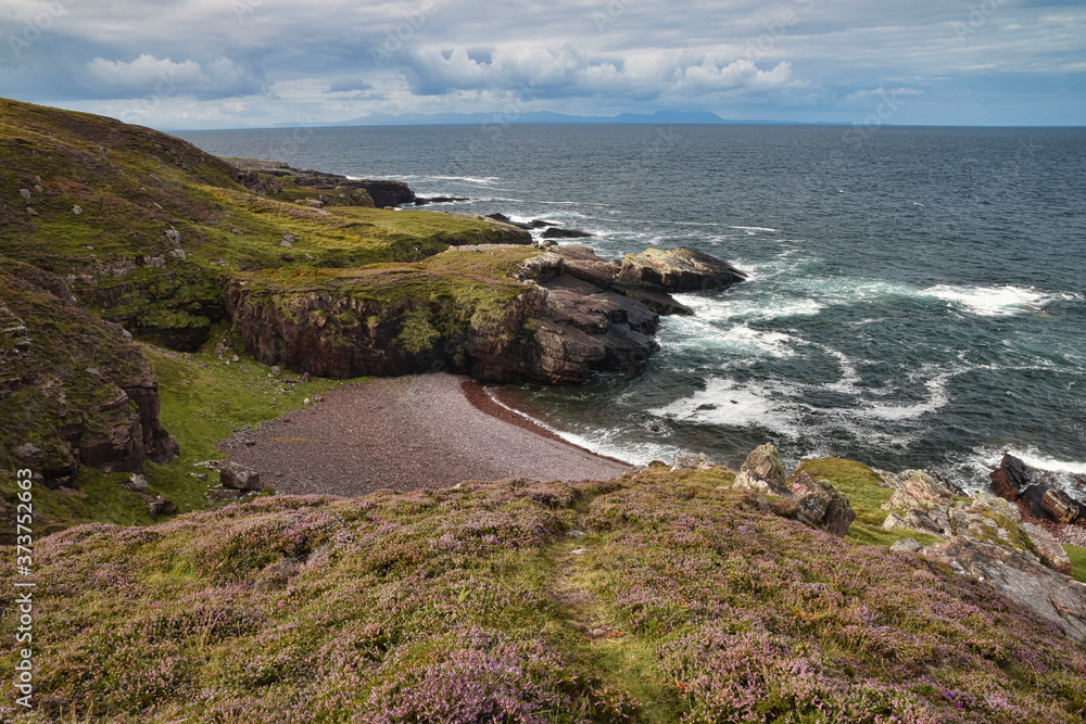 Lonely Scottish beach near Gairloch with heather and grassy path in foreground, pink shingle beach and cliffs, Isle of Skye in far distance.