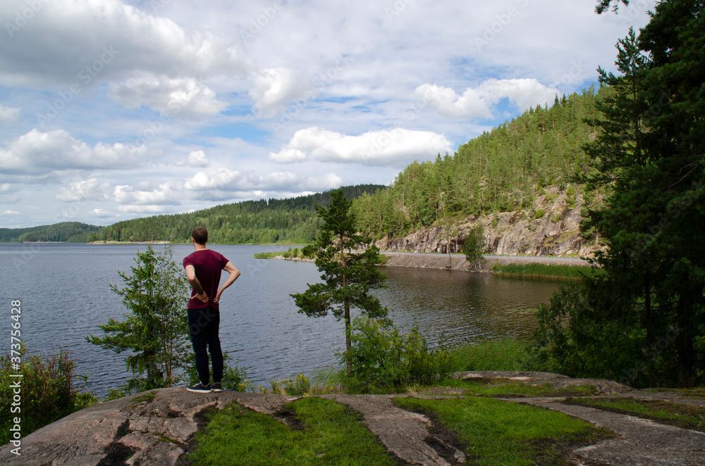 A young man stands admiring the beautiful views. Lake, fir trees on the hills, blue sky with clouds. Northern landscapes in Karelia, Russia.