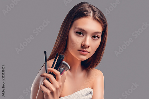 Cute attractive girl holding makeup brushes isolated on gray background. Skin care concept. Spa treatments, cosmetology, make-up.