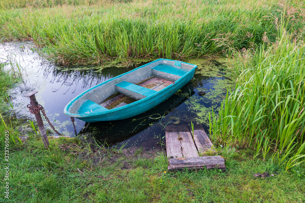 An old blue boat is tied with a rusty chain to a metal pole. The boat is standing in a small grassy pond. There are remains of a wooden bridge.