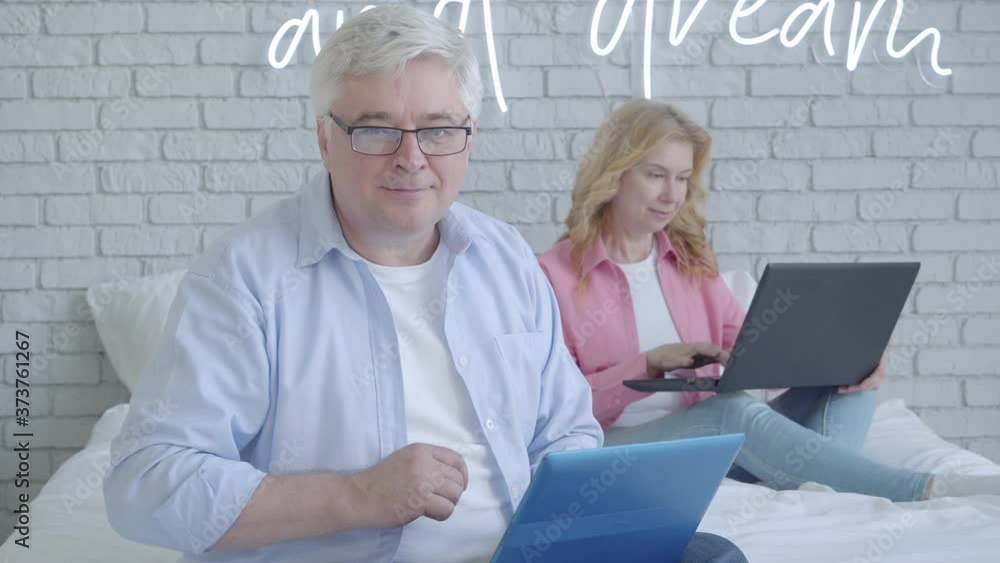 Handsome senior man typing on laptop keyboard, looking at camera and smiling. Blurred woman surfing Internet at the background. Portrait of happy mid-adult Caucasian man working online on Covid-19. Handsome senior man typing on laptop keyboard, looking at camera and smiling. Blurred woman surfing Internet at the background. Portrait of happy mid-adult Caucasian man working online on Covid-19.