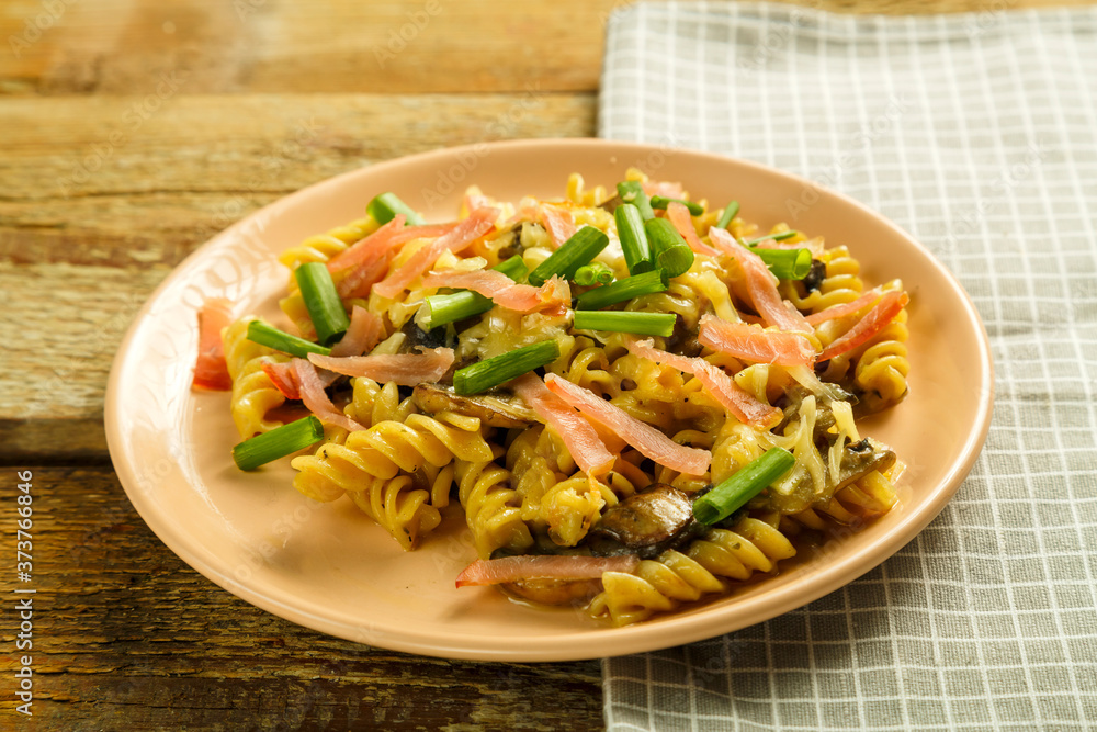 a plate with pasta with ham and mushrooms in a creamy sauce on the table with a fork on a napkin next to the cream. a plate with pasta with ham and mushrooms in a creamy sauce on the table with a fork on a napkin next to the cream.