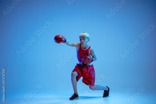 Teenager in sportswear boxing isolated on blue studio background in neon light. Novice male caucasian boxer training hard and working out, kicking. Sport, healthy lifestyle, movement concept.