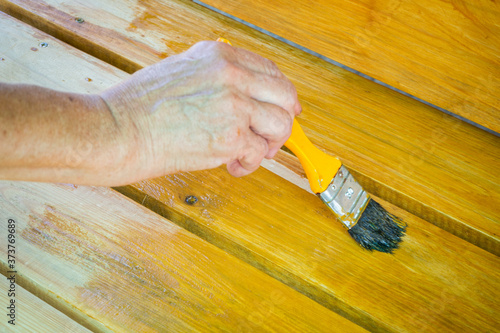 Painting a wooden bench with protective weather-resistant paint. The woman's hand applies a yellow impregnation with a brush. Surface treatment from insects. The concept of DIY. Close up