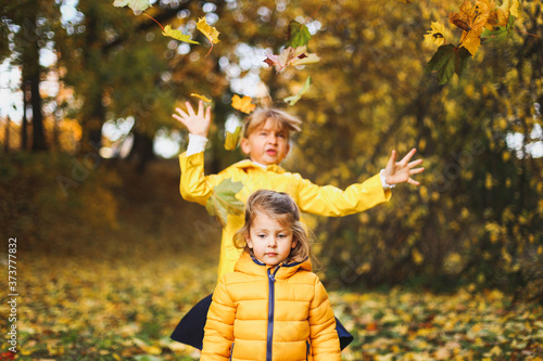 Little funny girls, sisters in stylish yellow raincoat, jacket play in autumn forest or park outdoors, throw up above head old maple leaves and foliage.Concept fall family photo shoot. Games,activity