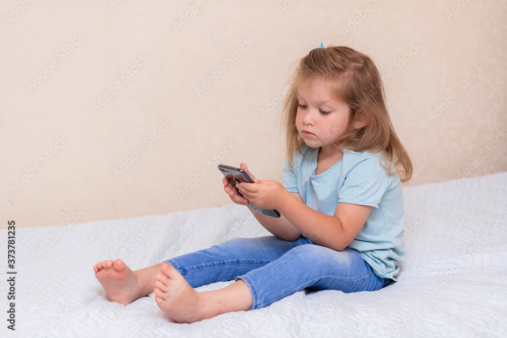 Curious little girl in blue clothes, uses phone applications,looking at a mobile device, sits on a bed.Clear girl, holds a smartphone, plays a game online alone at home.Concept of a child and a gadget Curious little girl in blue clothes, uses phone applications,looking at a mobile device, sits on a bed.Clear girl, holds a smartphone, plays a game online alone at home.Concept of a child and a gadget