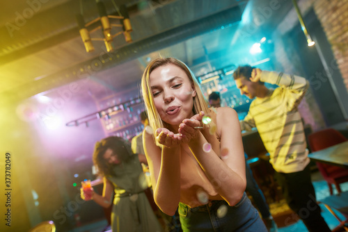 Dance and Party. Attractive caucasian young woman blowing confetti to camera while posing in the night club. Friends dancing, celebrating, having fun in the background