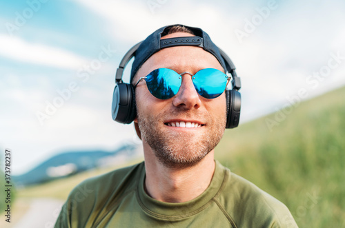 Cool man in a baseball cap, wireless headphones and blue sunglasses smiling and enjoying favorite music playing. © Soloviova Liudmyla