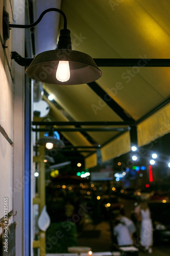 decorative black iron lantern in loft style with edison bulb on the wall of facade street cafe with a canopy, hanging street lighting glowe with warm light, night scene.
