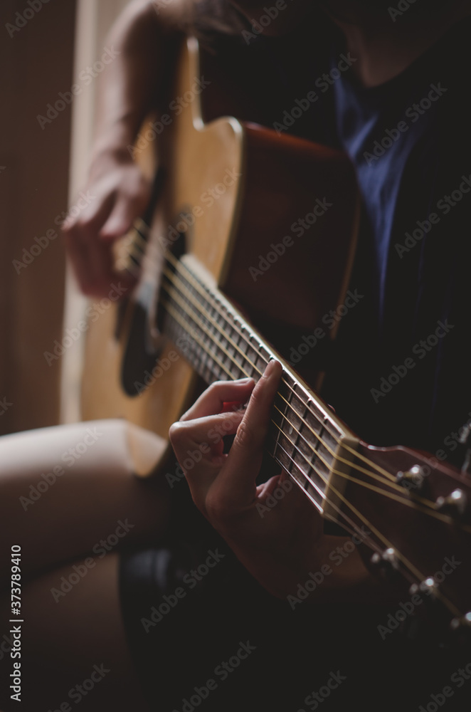 The girl plays the guitar in the room.Dim light from the window