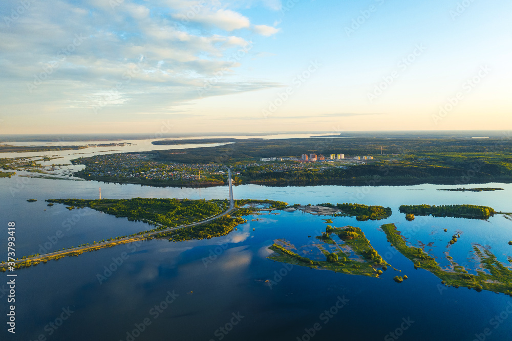 View from a drone, from a bird's-eye view of a wide blue river, islands on the river, bridge, city by river, sky with clouds, green forest.