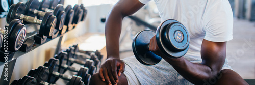 Foto Young African American man sitting and lifting a dumbbell close to the rack at gym