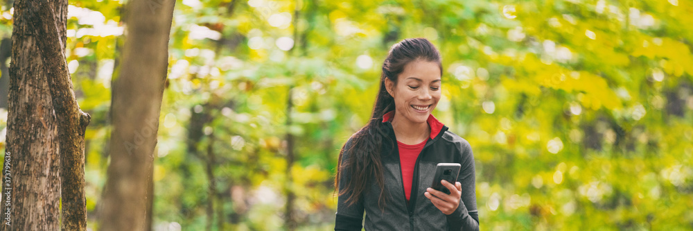 Autumn walk in the forest banner. Asian woman texting using mobile phone on outdoor hike walking in nature trail path panoramic background. Happy girl holding cellphone on active leisure activity. Autumn walk in the forest banner. Asian woman texting using mobile phone on outdoor hike walking in nature trail path panoramic background. Happy girl holding cellphone on active leisure activity.