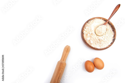 Baking ingredients: Wheat flour in a bowl or wooden plate, brown eggs and a rolling pin on a white background. The view from the top.