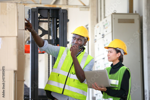 African American and Asian workers wearing safety vest while working in warehouse checking for the inventory of product using laptop 