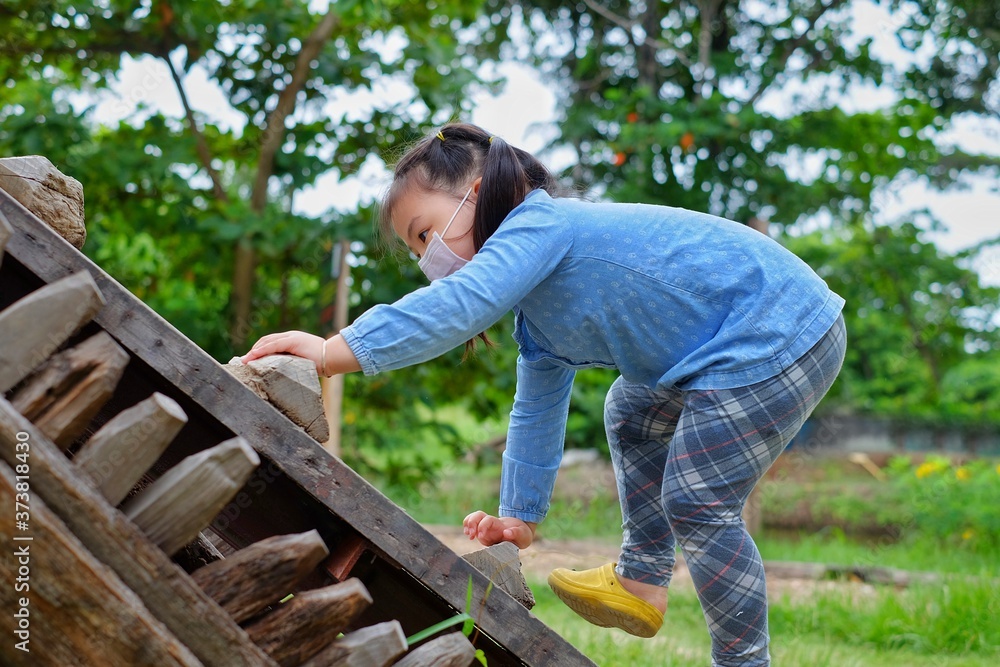 A cute young Asian girl wearing a surgical mask, climbing up a wooden steep ramp at a park, grabbing the steps with her hands, being brave but cautious. A cute young Asian girl wearing a surgical mask, climbing up a wooden steep ramp at a park, grabbing the steps with her hands, being brave but cautious.