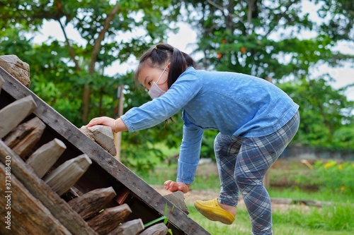 A cute young Asian girl wearing a surgical mask, climbing up a wooden steep ramp at a park, grabbing the steps with her hands, being brave but cautious.