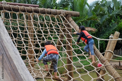 Two cute young Asian girls wearing orange life vest are going up a climbing net in an obstacle course at a summer camp.
