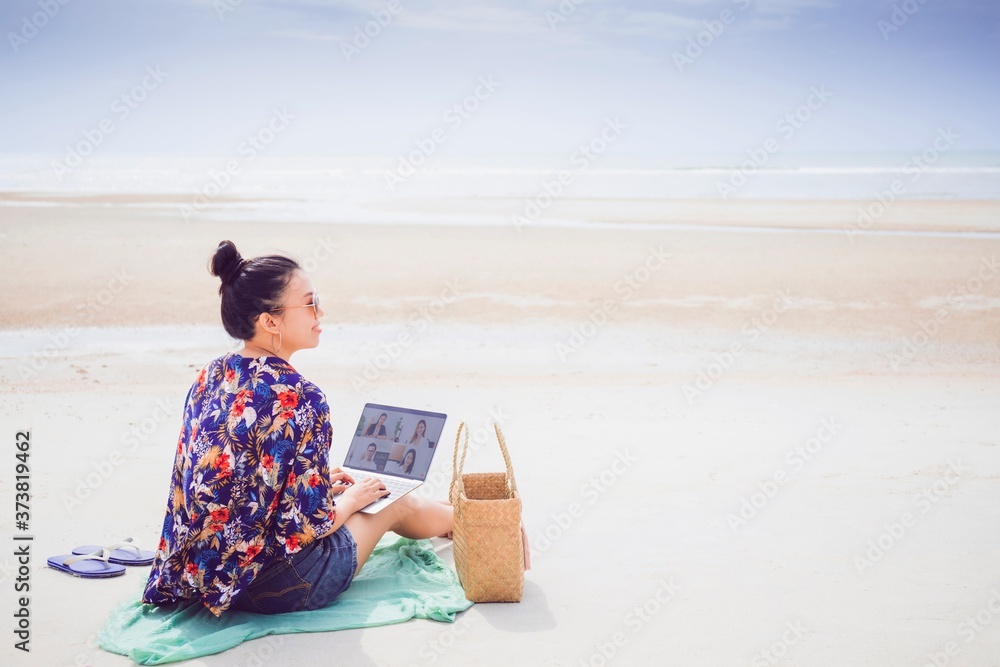 Asian happy woman sitting and using laptop meeting online on the beach by the sea background.Concept of working on vacation.