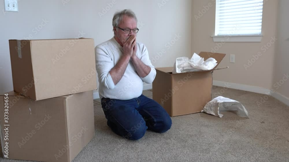 Distraught senior man in casual clothes sits among cardboard moving boxes. Runs hand through hair and looks sadly around apartment. Distraught senior man in casual clothes sits among cardboard moving boxes. Runs hand through hair and looks sadly around apartment.