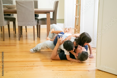 Laughing father lying on floor and hugging cute children. Happy attractive Caucasian dad embracing adorable kids, smiling and playing with son and daughter. Fatherhood, leisure and parenthood concept