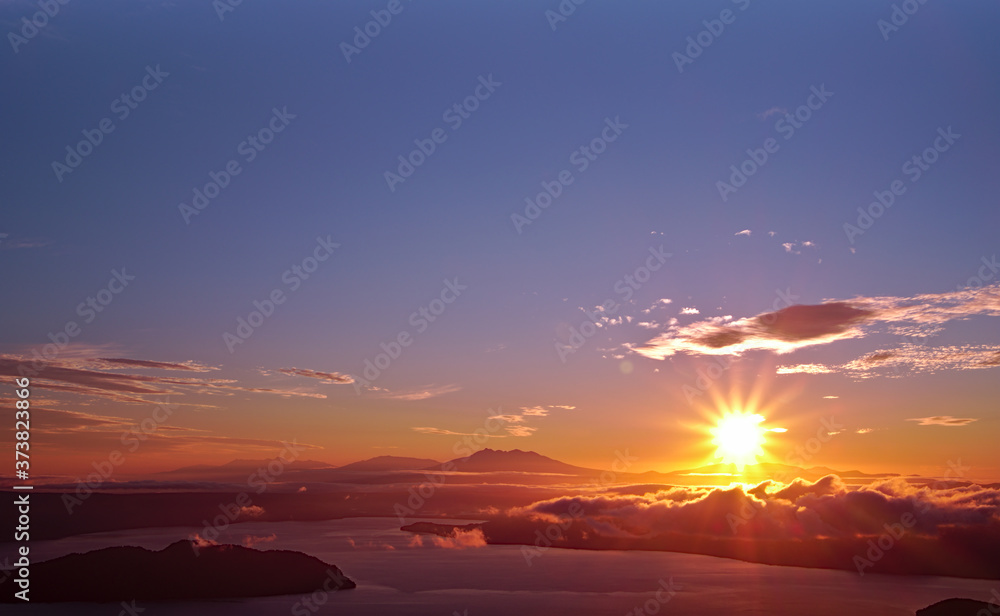 夜明けの空に力強く輝く太陽。津別峠、北海道。Abstract of sunrise and sunset. The sun is shining in the sky,clouds flowing low,lake surface seen in thin light.Scenic view from high place. Tsubetsu-pass,hokkaido,japan.