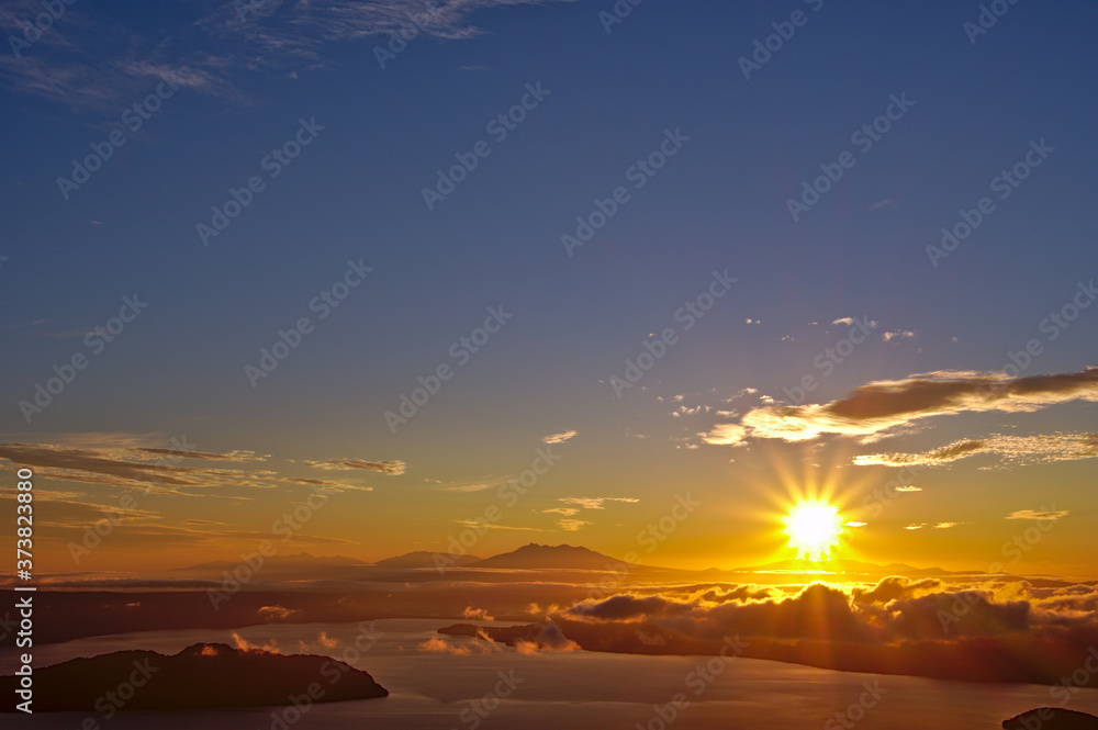 夜明けの空に力強く輝く太陽。津別峠、北海道。Abstract of sunrise and sunset. The sun is shining in the sky,clouds flowing low,lake surface seen in thin light.Scenic view from high place. Tsubetsu-pass,hokkaido,japan.