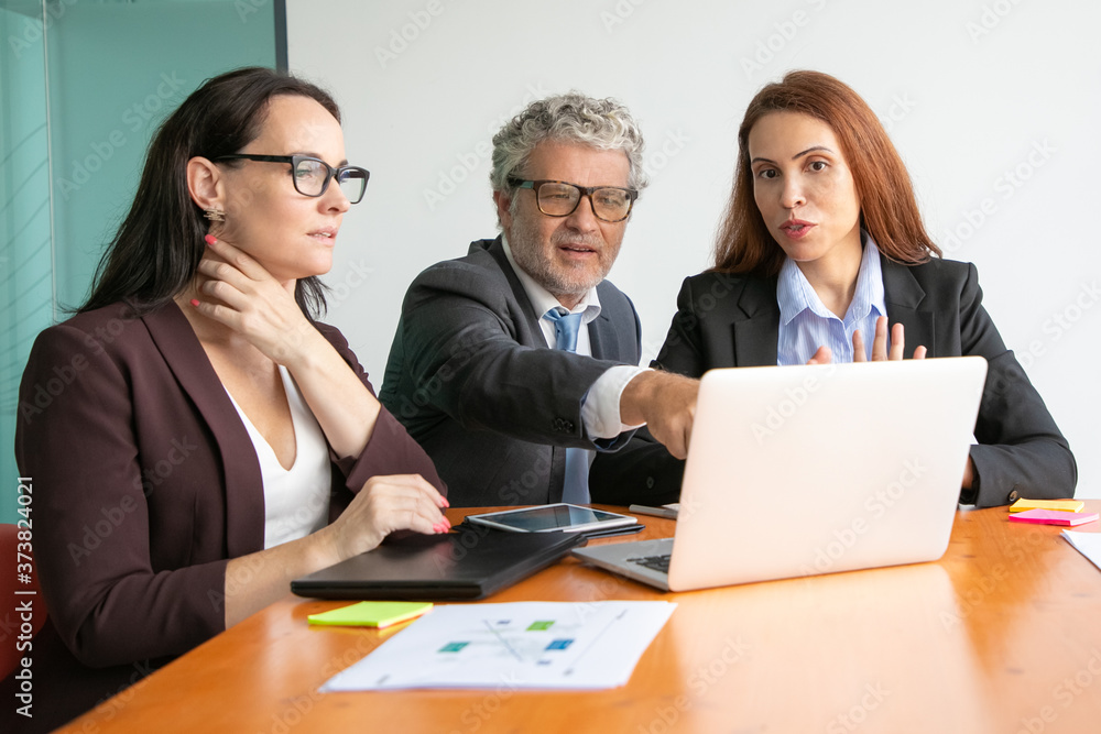Business people watching and discussing presentation on laptop, looking and pointing at display, sitting at meeting table with paper reports and charts. Medium shot. Marketing and teamwork concept