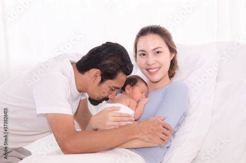 Happy Asian family lifestyle, father hold wife kissing toddler newborn baby with love, mother hug infant in arm sleeping comfort nursing medical care in bed with husband. baby sleep feeling safe
