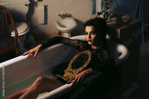 A woman in a black dress and with a mirror in her hands lies in the bath
