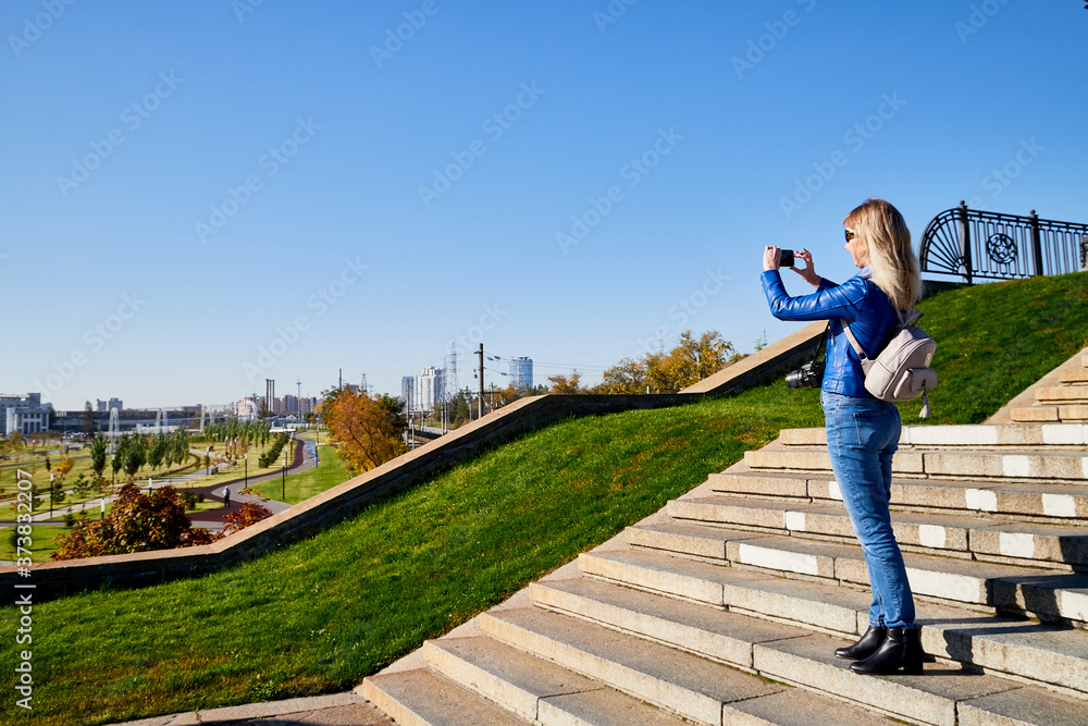 Attractive beautiful young girl holding cell phone in autumn park and making selfie. Traveller on the steps