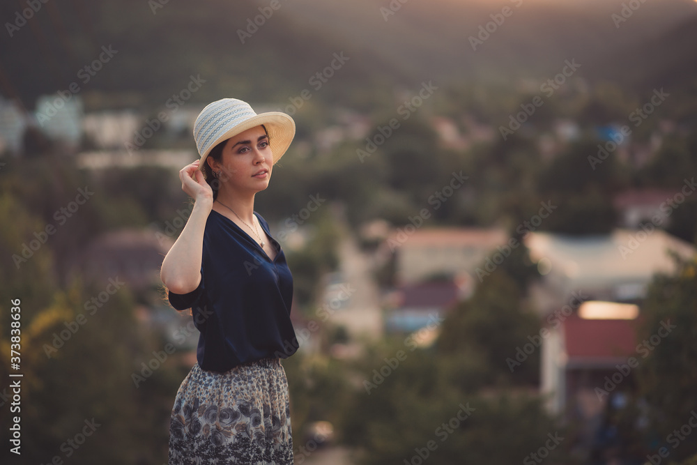 An elegant woman holds her hat in her hand and poses in a half-turn. Side view. In the background, a view of the city. Copy space. Travel and leisure concept