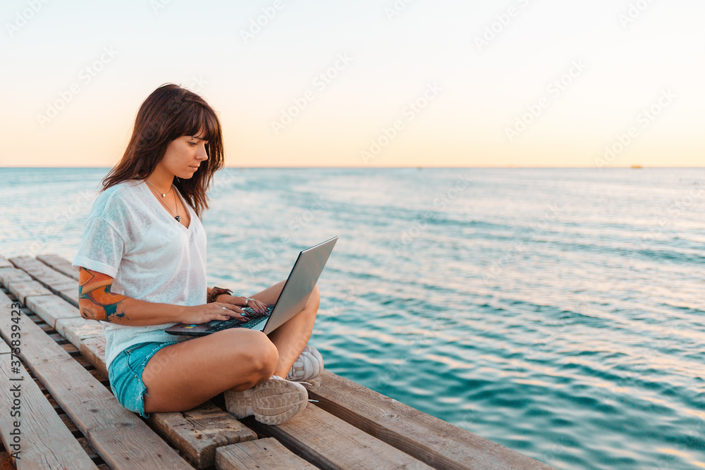 A young beautiful woman with tattoos is sitting on a pier by the sea and working at a laptop. Sea and sky in the background. Copy space. Concept of remote work and summer holidays