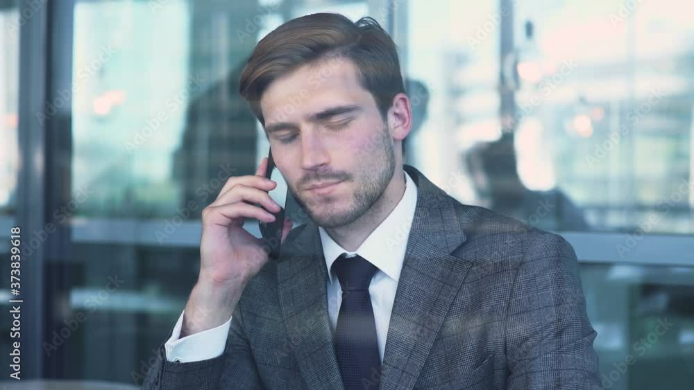 Young businessman in a suit is sitting on the terrace of a cafe and working on a laptop, talking on a mobile phone, view through the glass. Young businessman in a suit is sitting on the terrace of a cafe and working on a laptop, talking on a mobile phone, view through the glass.
