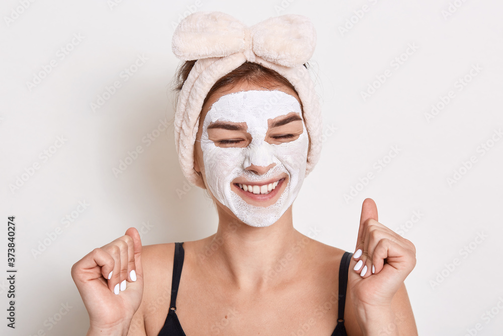 Photo of glad satisfied woman keeps eyes closed with toothy smile, has cosmetic procedures at home, applies clay mask for skin care, doing anti aging manipulation. Photo of glad satisfied woman keeps eyes closed with toothy smile, has cosmetic procedures at home, applies clay mask for skin care, doing anti aging manipulation.