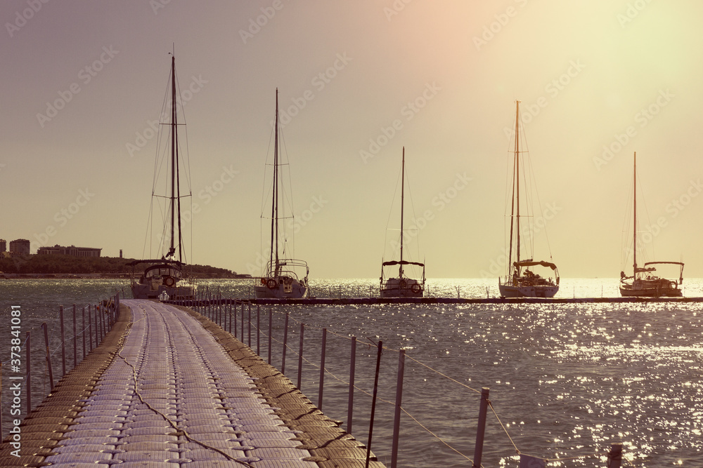 Several yachts in a row are parked at the pier. Sunset sky and sea in the background. Sunlight