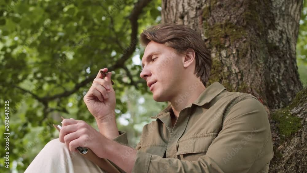 Pan shot of young Caucasian man with blond hair wearing casual clothes is sitting under the tree in woods and writing in notepad Pan shot of young Caucasian man with blond hair wearing casual clothes is sitting under the tree in woods and writing in notepad