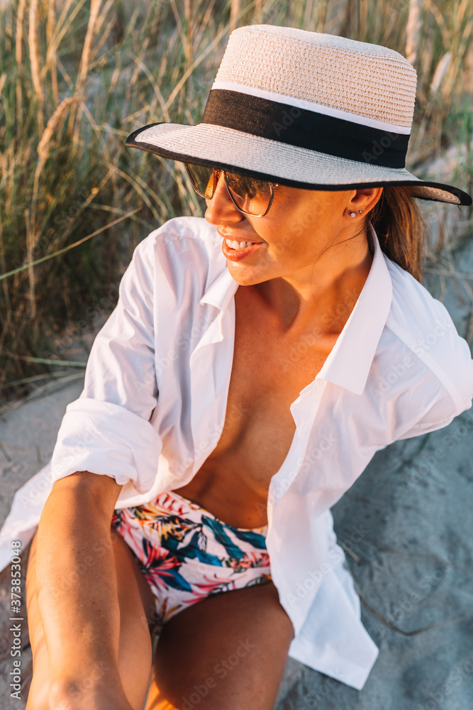 Beautiful boho styled model wearing white shirt and hat posing on the beach in sunlight. Tan, travel, vacation concept.