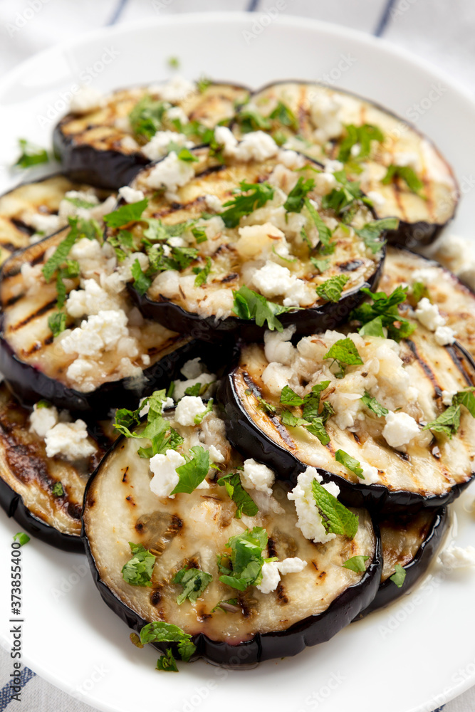 Homemade Grilled Eggplant with Feta and Herbs on a white plate on cloth, side view. Homemade Grilled Eggplant with Feta and Herbs on a white plate on cloth, side view.
