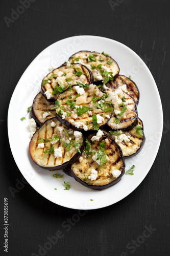 Homemade Grilled Eggplant with Feta and Herbs on a white plate on a black surface, high angle view.