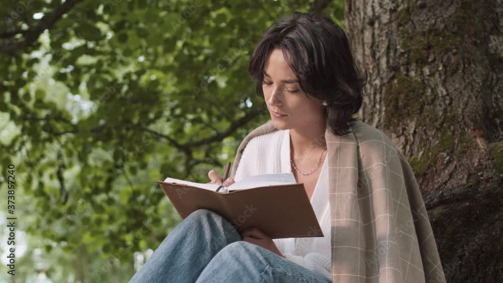 Lockdown of young pretty woman wearing casual clothes is sitting under the tree in park, writing in notebook then looking at camera and smiling Lockdown of young pretty woman wearing casual clothes is sitting under the tree in park, writing in notebook then looking at camera and smiling